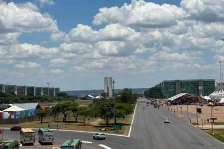 Imagem colorida da Esplanada dos Ministérios, em dia ensolarado. Há poucos carros na via, alguns ônibus e estruturas em montagem no gramado central