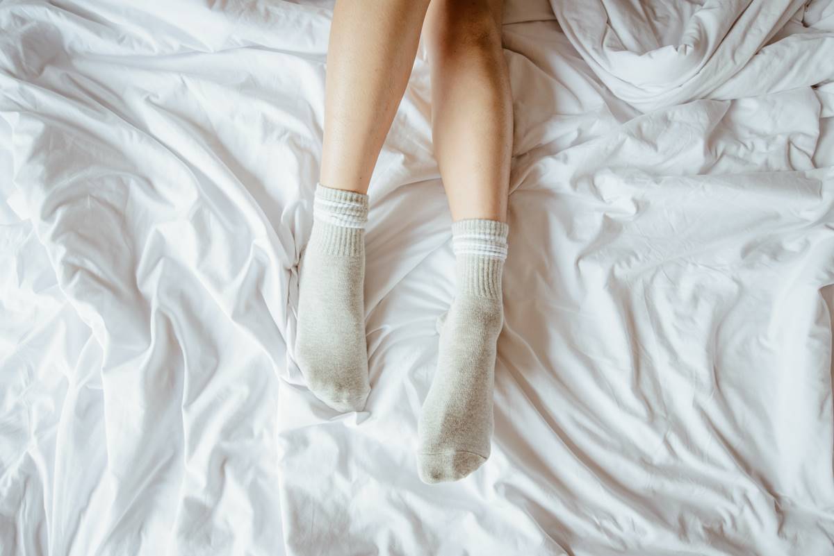 Foto colorida de dois pés com meias cinzas em cima de uma cama com lençol branco amassado - Metrópoles