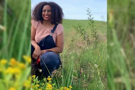 Foto colorida de mulher negra em meio a um campo verde com céu azul