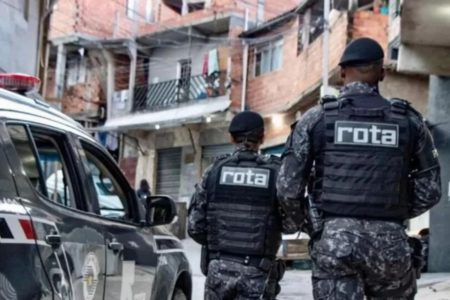 Imagem colorida de dois PMs da Rota, de costas, ao lado de viatura. Ao fundo, aparecem moradias simples, onde eles buscam suspeitos; Operação Escudo - Metrópoles