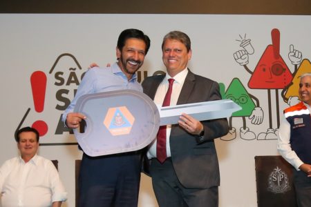 Imagem colorida mostra Ricardo Nunes e Tarcísio de Freitas, homens brancos de meia idade, segurando uma chave gigante simbólica de carro, feita de papelão, em evento de entrega de viaturas para a defesa civil