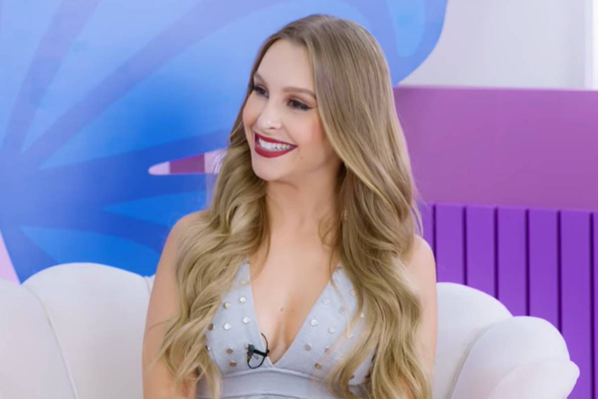 Foto colorida de um frame com a atriz Carla Diaz sentada de cabelos soltos, vestido cinza e batom vermelho - Metrópoles