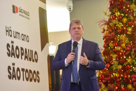 Imagem colorida mostra Tarcísio de Freitas em um palco, falando ao microfone, com árvore de natal ao fundo