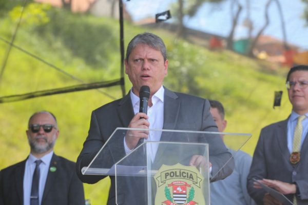 Imagem colorida mostra Tarcísio de Freitas, homem branco, de terno cinza e camisa branca, falando em um púlpito de acrílico com o logotipo da Polícia Civil, em um palco, com pessoas de roupas sociais atrás dele, e um fundo verde - Metrópoles