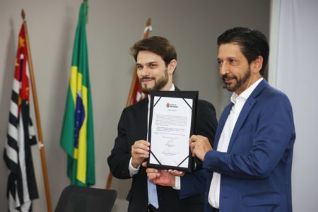 Imagem colorida mostra Felipe Becari, homem branco, jovem de cabelo e barba castanhos e aparados, segurando um documento com Ricardo Nunes, homem branco, de cabelo e barba pretos, de terno azul - Metrópoles