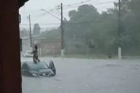 Imagem colorida mostra mulher em cima de um carro no meio de uma rua alagada em São Paulo durante chuva - Metrópoles