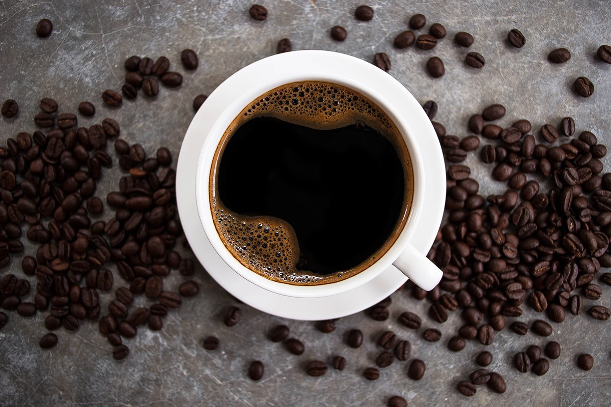 Café preto em um copo branco colocado sobre uma mesa de cimento com grãos de café - Metrópoles