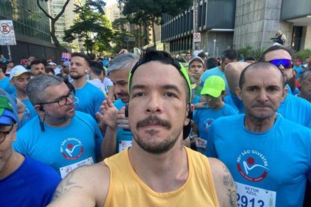 Em foto colorida homem branco de barba e camiseta regata laranja clara em meio a homens com camisetas azuis claras da São Silvestre - Metrópoles