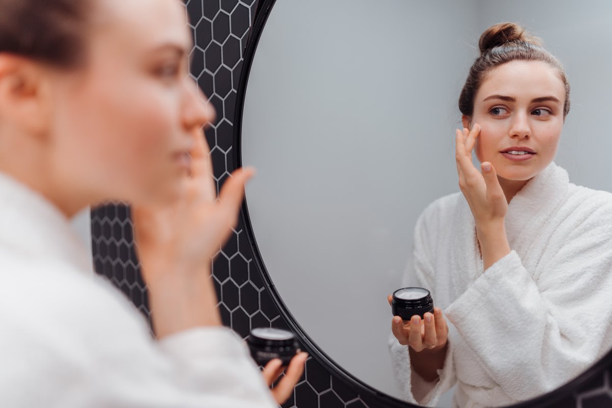 Mulher branca com cabelo preso no alto da cabeça aplicando um creme no rosto; ela usa um roupão branco