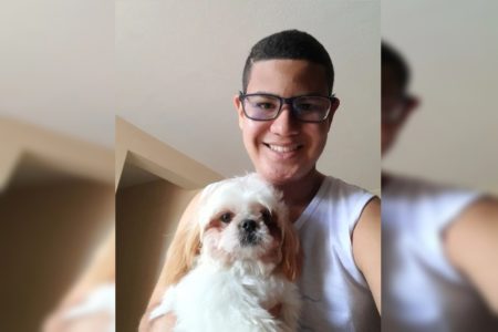 Foto de jovem com óculos, camisa branca e cabelo curto. Ele está com cachorrinho no colo