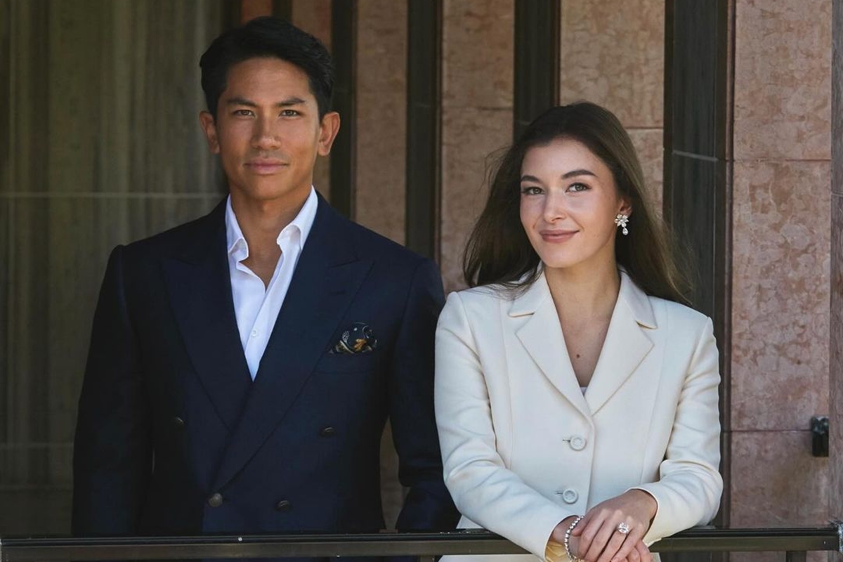 Foto de homem, vestido com blazer azul e blusa branca, ao lado de mulher com blazer nude. Ela está mostrando um anel. Os dois estão em uma varanda - Metrópoles