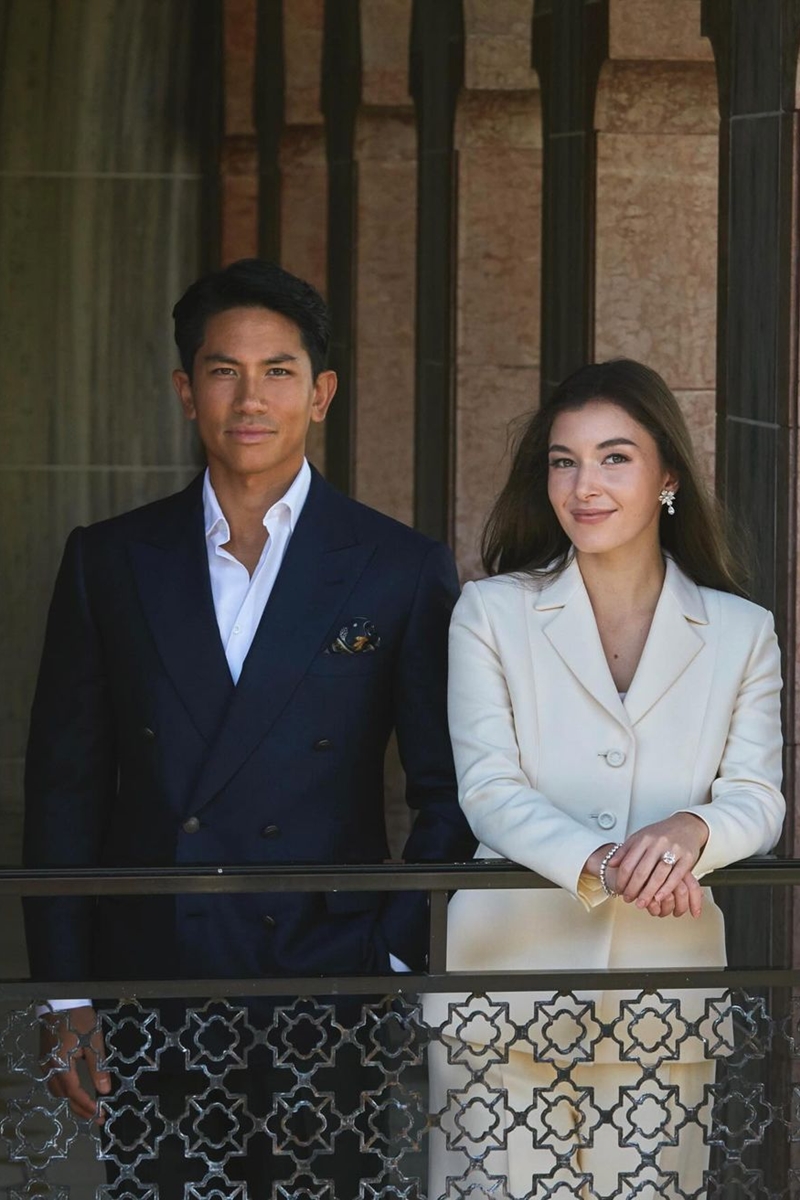 Foto de homem, vestido com blazer azul e blusa branca, ao lado de mulher com blazer nude. Ela está mostrando um anel. Os dois estão em uma varanda - Metrópoles