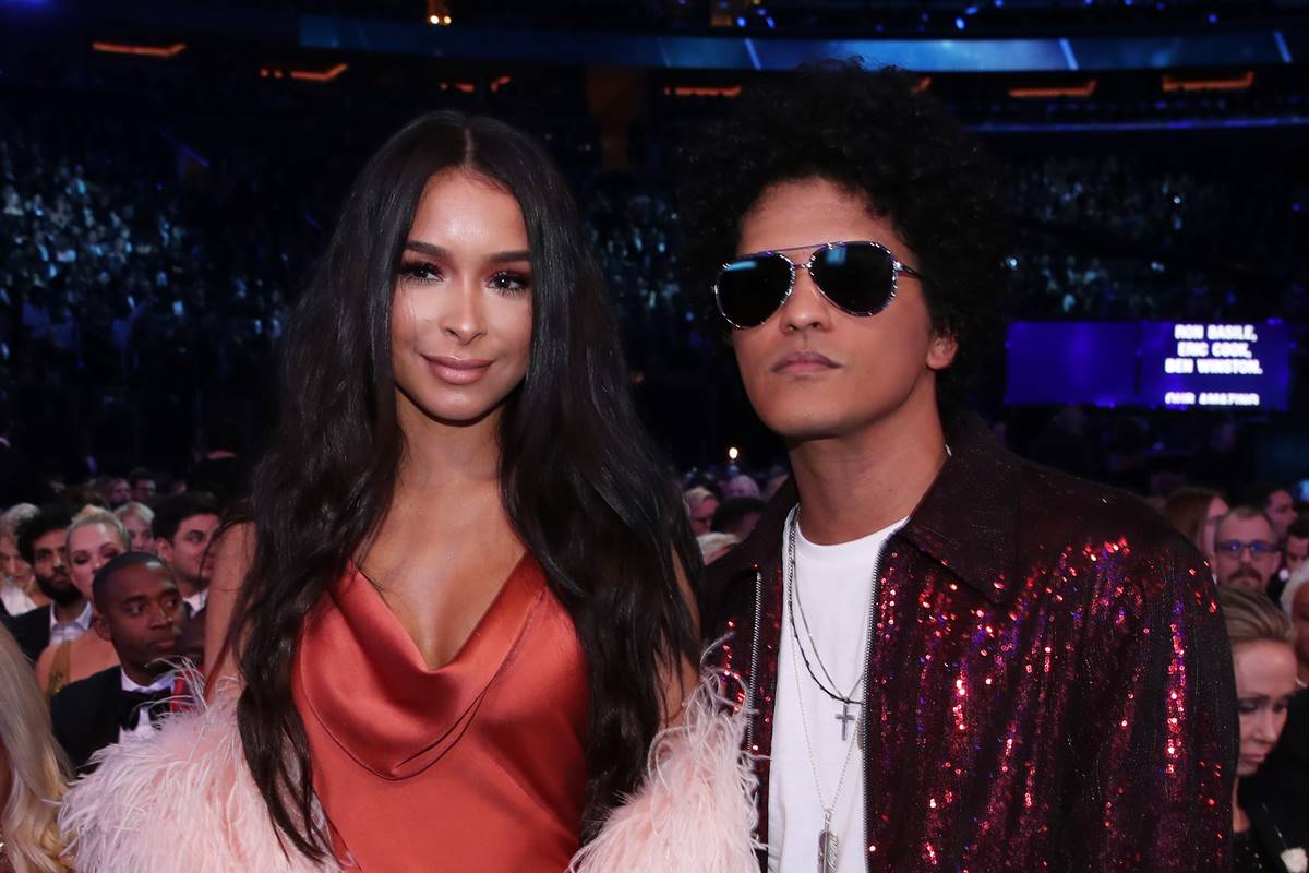 Bruno Mars e Jessica Caban em evento, usando roupas sociais. Cantor aparece de óculos escuros - Metrópoles