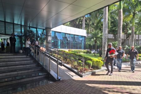 Imagem colorida mostra a entrada de um velório, com escadaria e rampa para cadeirantes, e Alexandre Frota, homem branco vestindo roupas pretas e trazendo um embrulho vermelho, se direcionando à entrada - Metrópoles