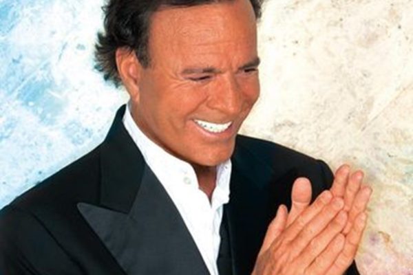 Julio Iglesias posa, sorridente, com as mãos juntas e blazer preto - Metrópoles