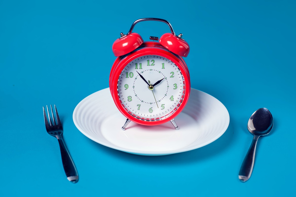 Prato de comida branco com relógio vermelho em cima, talheres ao lado e fundo azul - Metrópoles