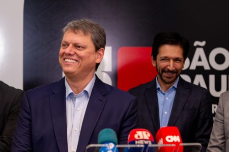 Imagem colorida mostra Tarcísio de Freitas, homem branco, grisalho, de terno azul marinho e camisa azul clara, com um púlpito de microfones à sua frente. Atrás dele, está Ricardo Nunes, homem branco, de cabelo e barba pretos, vestindo terno e camisa. Ambos sorriem na foto, da cintura para cima - Metrópoles
