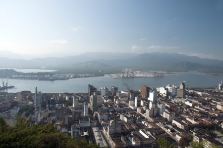 Imagem colorida mostra vista aérea do porto de Santos, com o canal no meio e a cidade do Guarujá ao fundo - Metrópoles