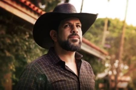 Foto de um homem branco, com barba preta, usando chapéu marrom e blusa quadriculada marrom. Ele está em uma fazenda - Metrópoles