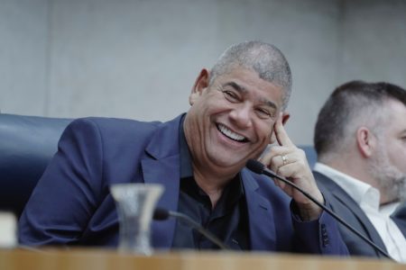 Imagem colorida mostra Milton Leite, homem negro, de terno azul e camisa preta, ao lado de um microfone em uma mesa. O presidente da Câmara de vereadores sorri - Metrópoles