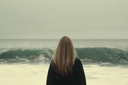 Mulher loira de costas com casaco contemplando as ondas - Metrópoles