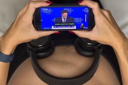 Fotografia colorida mostra barriga de Marina Helena, que está grávida, com fones de ouvido posicionados sobre ela e o discurso de Javier Milei transmitido em tela de celular - Metrópoles