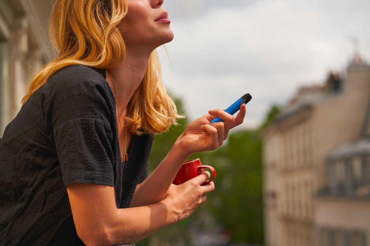 Fotografia colorida mostrando mulher segurando caneca e vape, cigarro eletrônico na varanda-Metrópoles