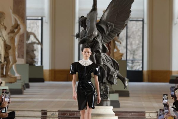 Com estátua escultural ao fundo, modelo desfile com vestido preto de vinil, que tem pérolas na gola alta, com mandas curtas bufantes - Metrópoles