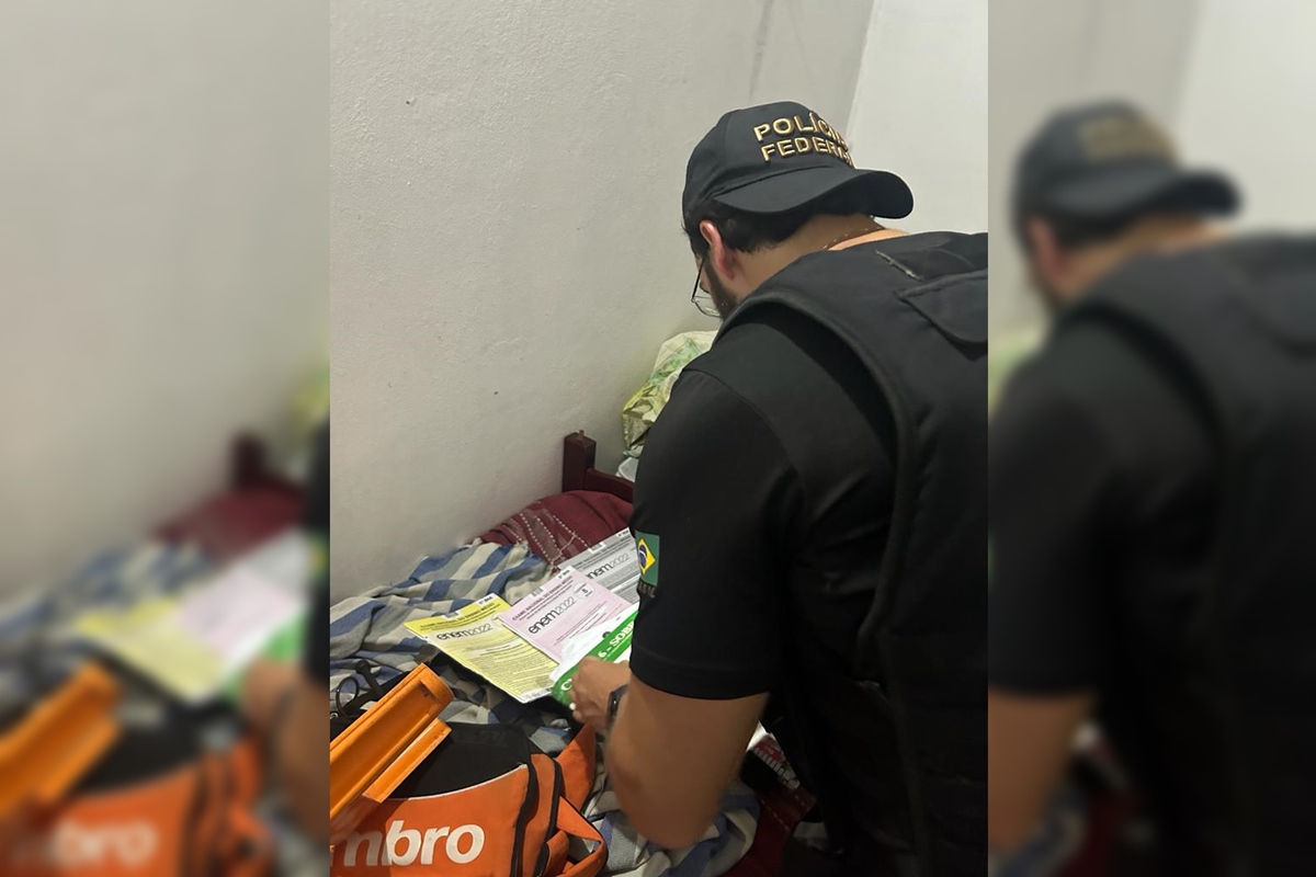 Imagem colorida de policial federal de costas, mexendo em objetos sobre uma mesa. Ele usa boné, colete e roupa pretos