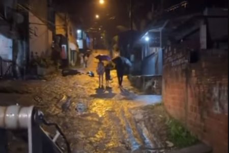 Foto colorida mostra Vila Sahy durante chuva forte. Duas pessoas aparecem caminhando com guarda chuva em viela cheia de lama