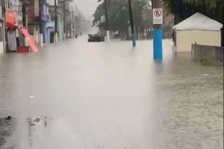 Imagem colorida mostra rua alada durante chuva. Água bate a altura de quase 30 centímetros em poste de trânsito