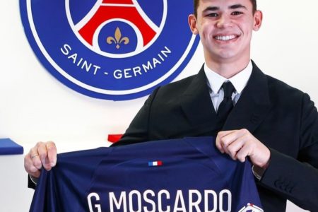 Gabriel Moscardo está vestindo terno em frente ao escudo do Paris Saint German e segura em suas maõs a camisa do PSG com seu nome nas costas