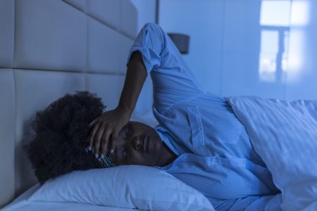 Imagem mostra mulher negra deitada de pijama na cama com a mão na cabeça - Metrópoles - dormir