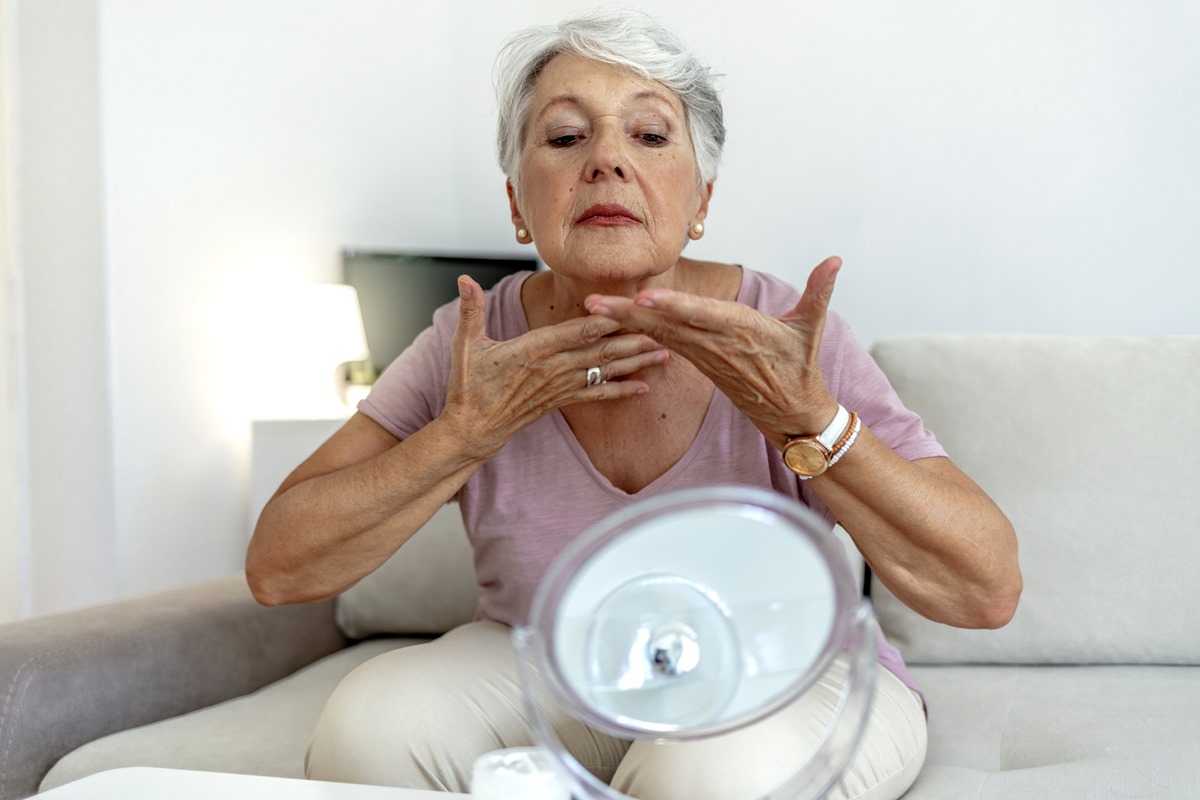 Foto de mulher madura, fazendo massagem facial no pescoço. Ela está se olhando no espelho e usa uma blusa rosé - Metrópoles