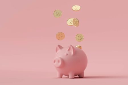 Foto colorida de um cofrinho de porquinho em um fundo cor de rosa com moedas douradas caindo em cima - Metrópoles
