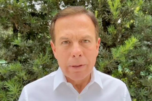 Imagem colorida mostra João Doria, homem branco, de cabelos pretos, camisa branca, em foto do peito para cima, na frente de um fundo de vegetação verde - Metrópoles