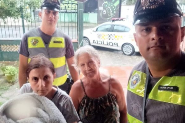 Policiais militares fardados de pé juntamente com avó e mãe de bebê enrolada em uma coberta