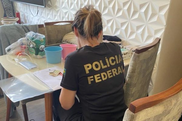 Mulher com camiseta da Polícia Federal (PF) sentada em cadeira averigua material apreendido em residência