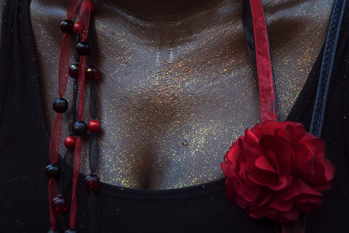 Foto colorida em close do colo de uma mulher suada com glitter na pele, uma blusa preta e uma rosa vermelha pendurada - Metrópoles