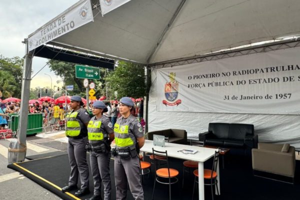 Três policiais militares fardadas estão de pé em frente a uma tenda de atendimento à vítimas de assédio