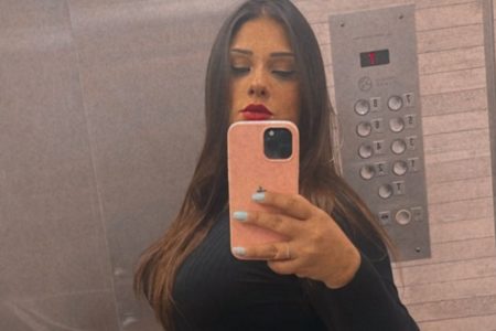 Mulher branca com cabelos escuros, batom vermelho e blusa preta segura celular em frente ao rosto. É empresária