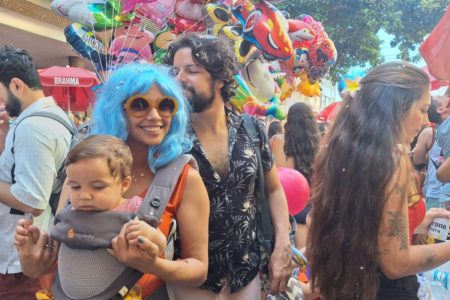 Foto colorida de mulher com peruca azul e óculos escuro carregando um bebê com diversas pessoas ao fundo durante bloco de Carnaval em São Paulo - Metrópoles