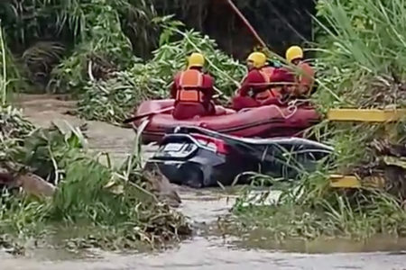Bote vermelho do Corpo de Bombeiros com três pessoas a bordo em córrego. Carro preto está parciamente submerso