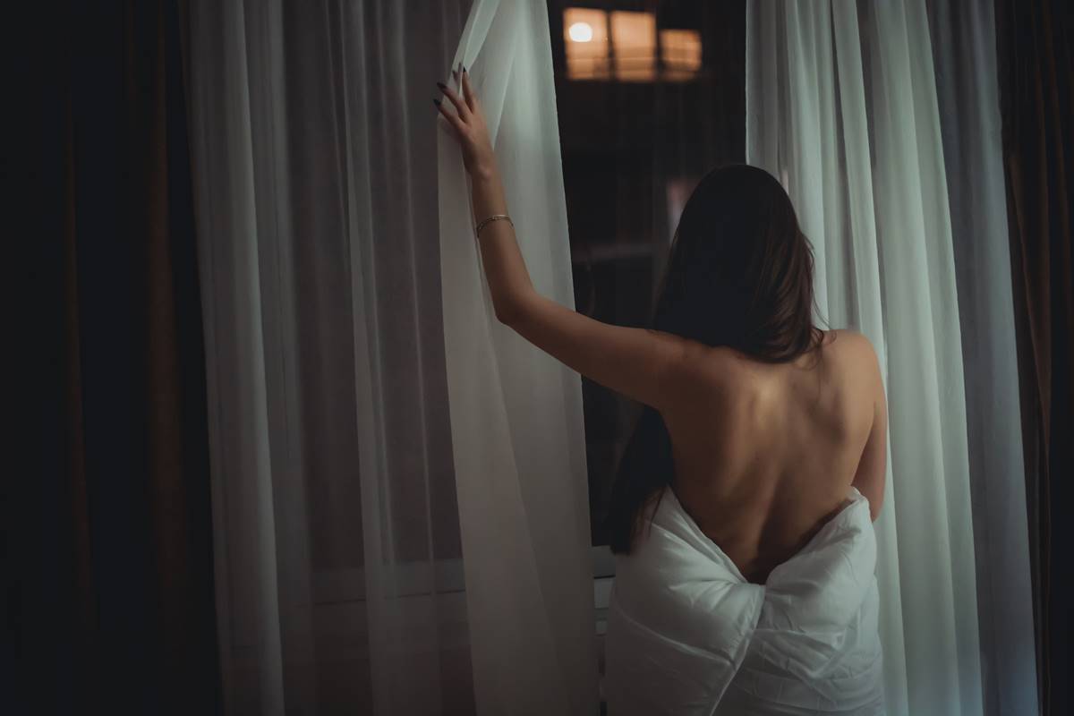 Foto colorida de uma mulher nua enrolada em um cobertor de costas olhando pela janela - Metrópoles