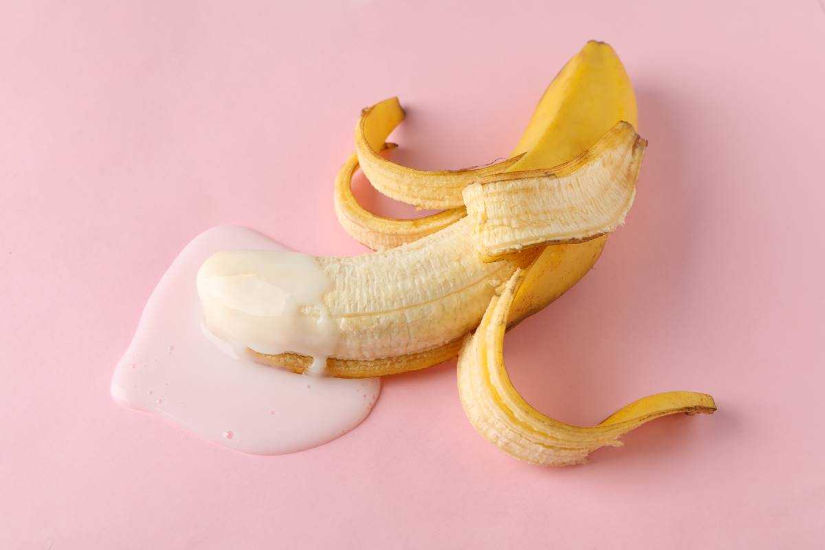 Foto colorida de uma banana meio descascada com um liquido branco na ponta em um fundo cor-de-rosa - Metrópoles