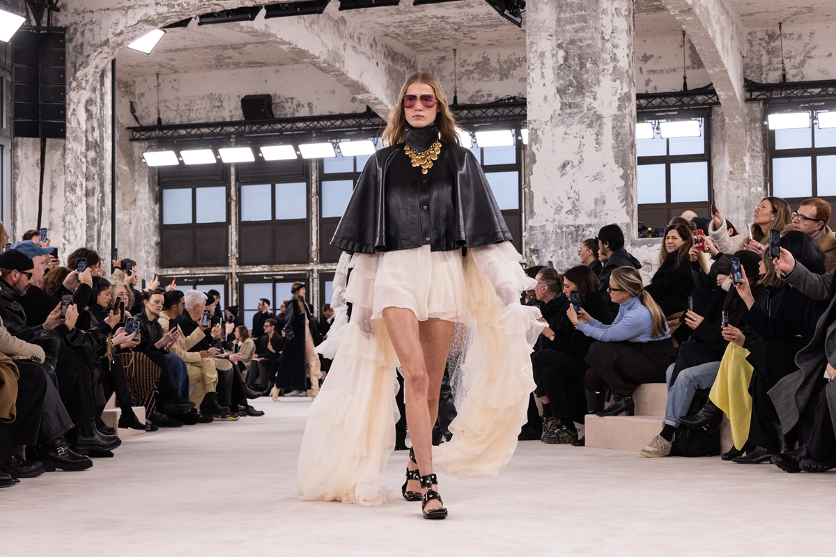 Modelo usa capa preta curta por cima de vestido creme com mangas esvoaçantes. Nos pés, sapatilha preta - Metrópoles