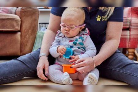 Foto mostra a bebê Margot, que nasceu sem olhos, usando um óculos protetor rosa e brincando com objetos coloridos enquanto é abraçada por um braços de uma mulher - Metrópoles