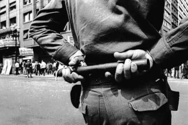 Imagem em preto e branco de policial fardado, de costas, segurando um cassetete. Ao fundo, manifestantes contra a ditadura militar -- Metrópoles