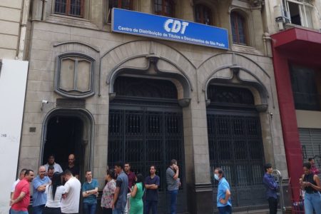 Imagem colorida de pessoas esperando na porta de um prédio. Metrópoles