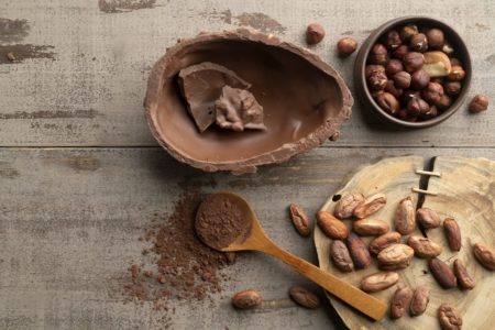 Foto mostra uma metade de um ovo de chocolate, pedaços de cacau e uma colher de pau com chocolate em pó em cima de uma mesa de madeira - Metrópoles
